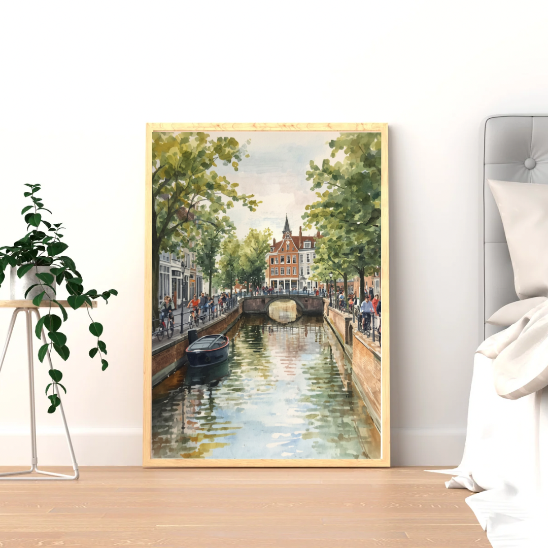 Amsterdam zomergracht schilderij in donkere lijst staand op lichte houten vloer tegen witte muur naast groene plant en decoratieve mand – aquarel wall art woonkamer
