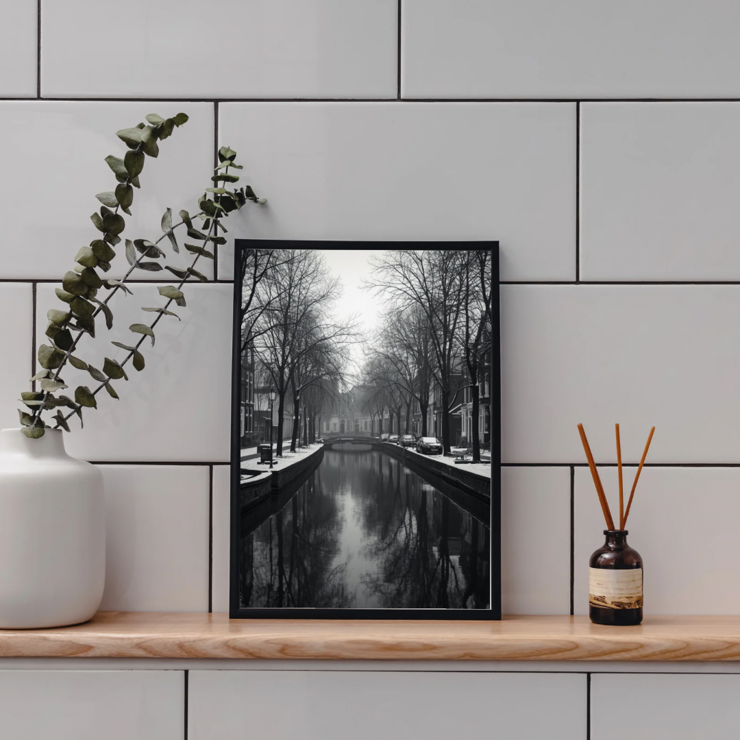 Alkmaar wintergracht schilderij in zwarte lijst aan witte tegelmuur boven houten plank met witte vaas en rieten diffuser in badkamer – zwart-wit wall art print
