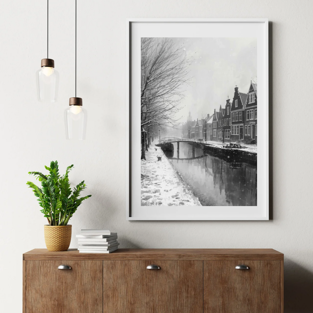 Alkmaar sneeuwval gracht schilderij in witte lijst aan witte muur boven donker houten dressoir met groene plant, boeken en hanglamp in hal – zwart-wit wall art print
