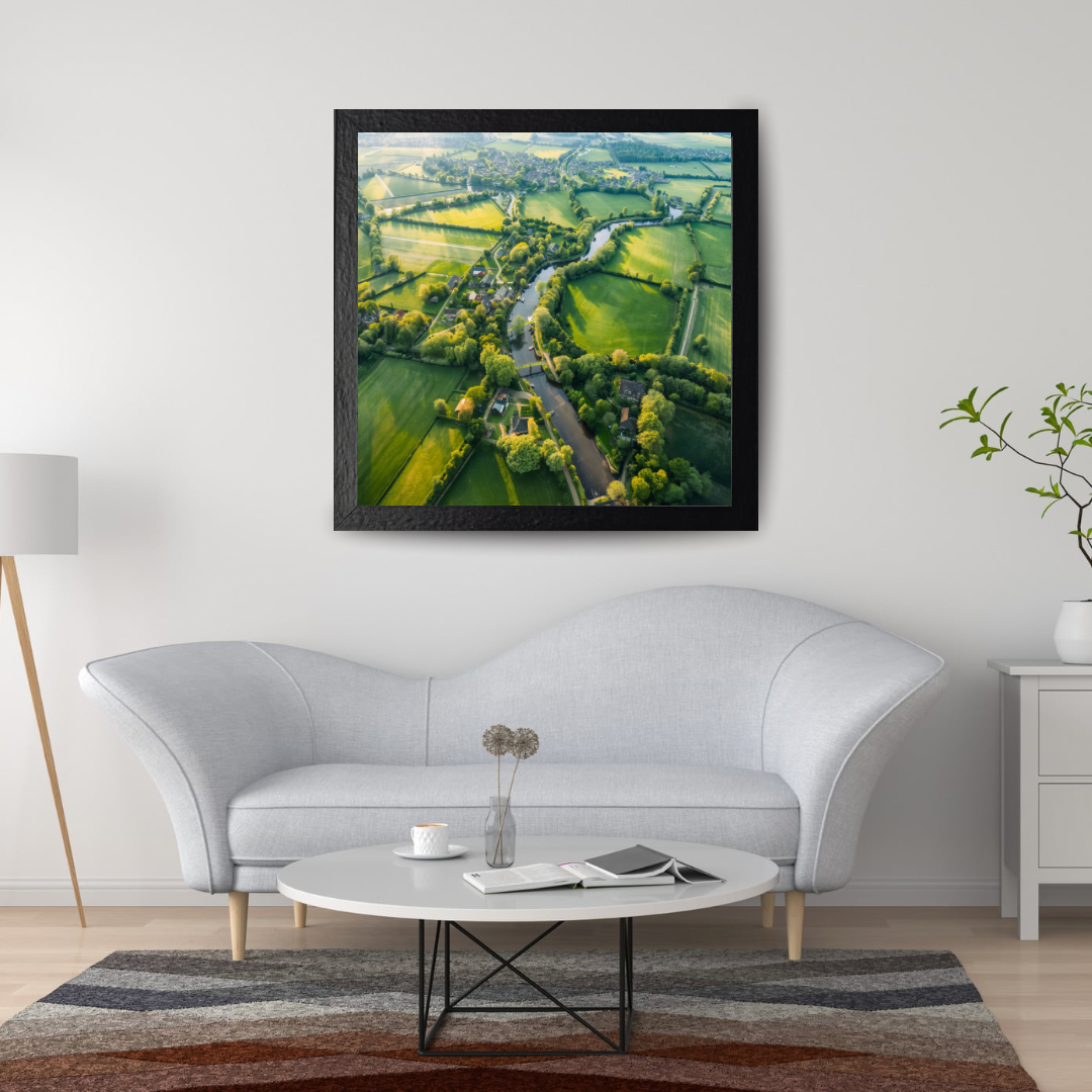 Weelderig groen landschap luchtfoto schilderij in zwarte lijst aan witte muur boven witte gebogen bank met ronde glazen salontafel en geometrisch vloerkleed – wall art print woonkamer
