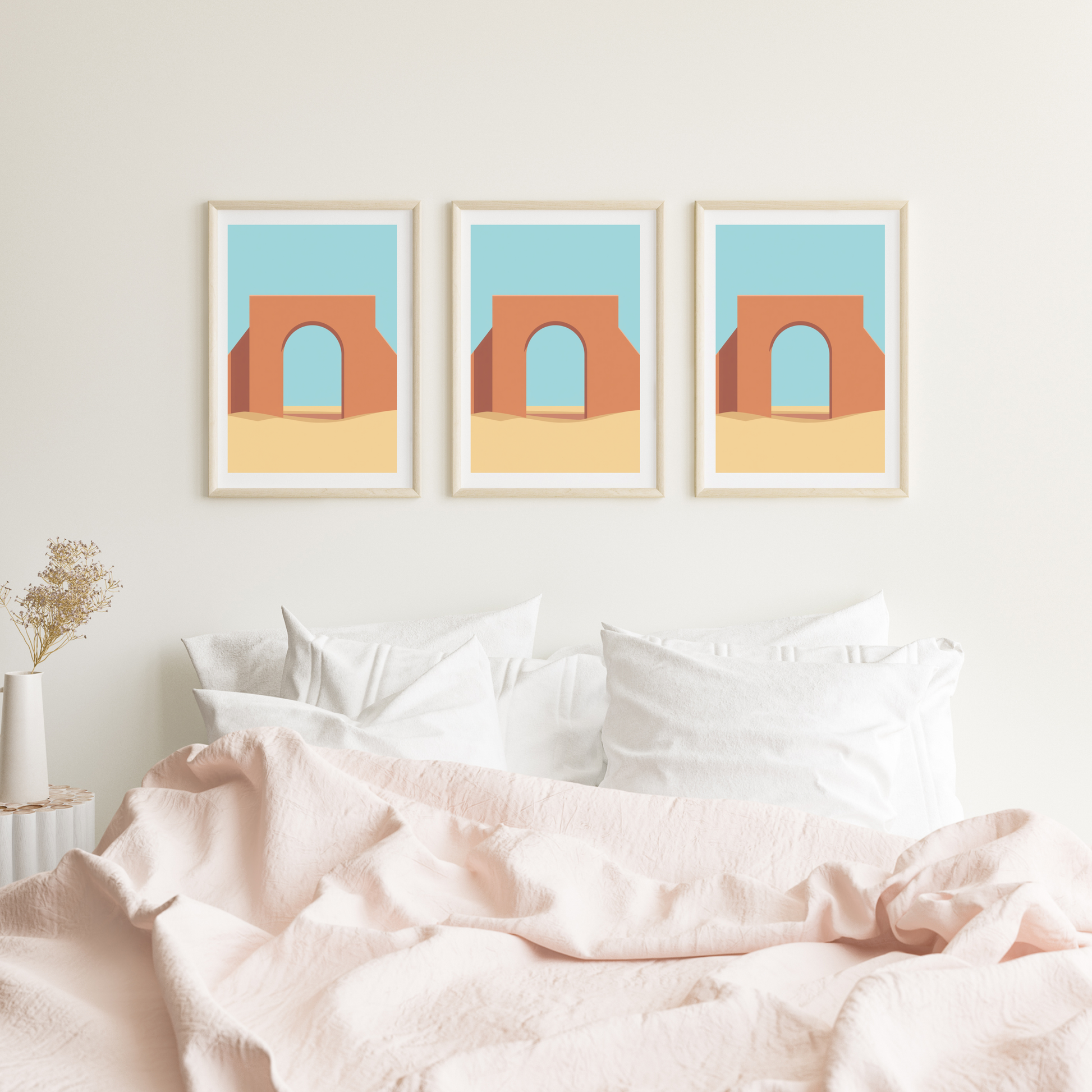 Marokkaanse poort schilderij met terracotta boog en turquoise lucht in drie witte lijsten naast elkaar hangend aan witte muur boven bed met roze beddengoed – mediterraan wall art logeerkamer
