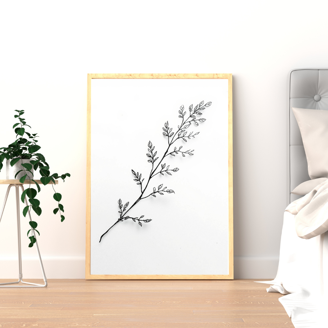 Botanische kruidentak schilderij met fijne zwarte kruidentak en delicate blaadjes in gouden lijst staand op houten vloer naast groene plant – minimalistisch wall art logeerkamer
