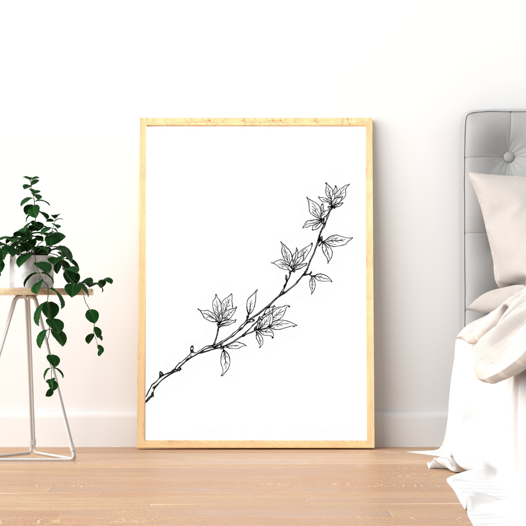 Botanische magnoliatak schilderij met fijne zwarte magnoliatak en witte bloesems in gouden lijst staand op houten vloer naast groene plant – elegant wall art logeerkamer
