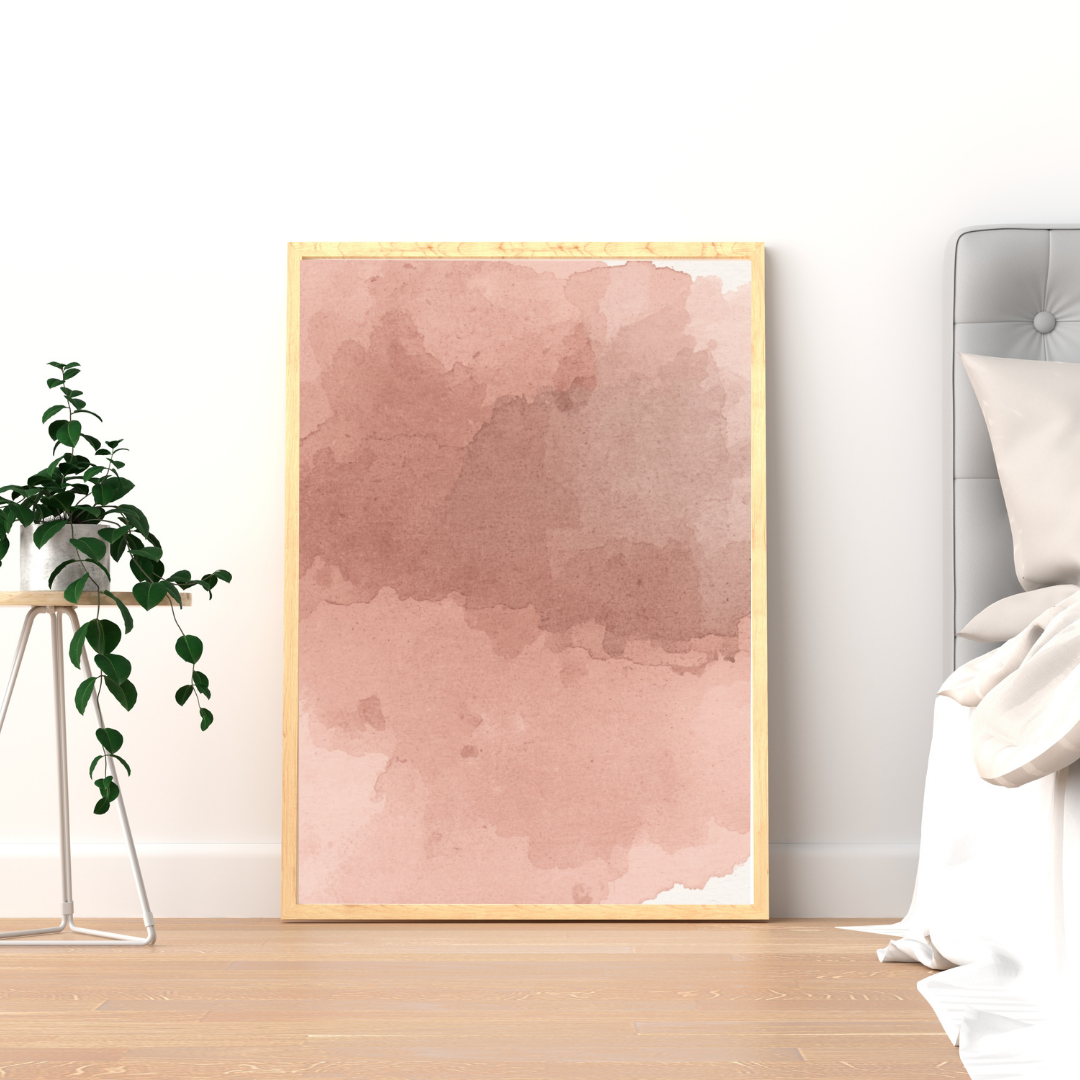 Abstract roze aquarel schilderij met zachte roze wolkenwassing in gouden lijst staand op houten vloer naast groene plant – dromerig wall art logeerkamer
